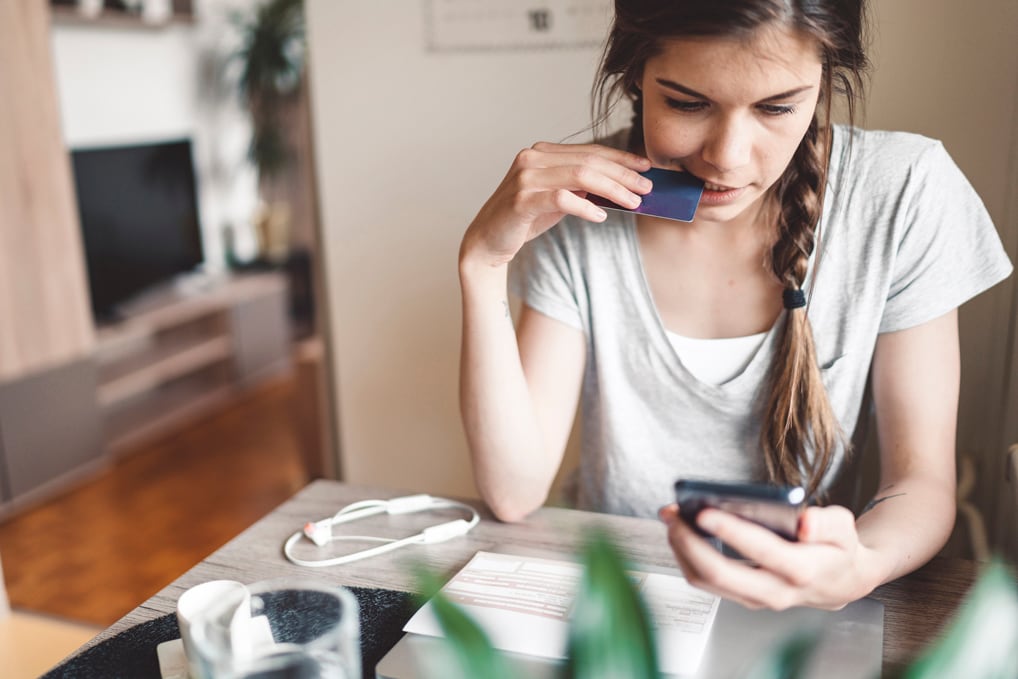 A young woman sitting at a table, holding a credit card in one hand and a looking at smartphone in the other hand.