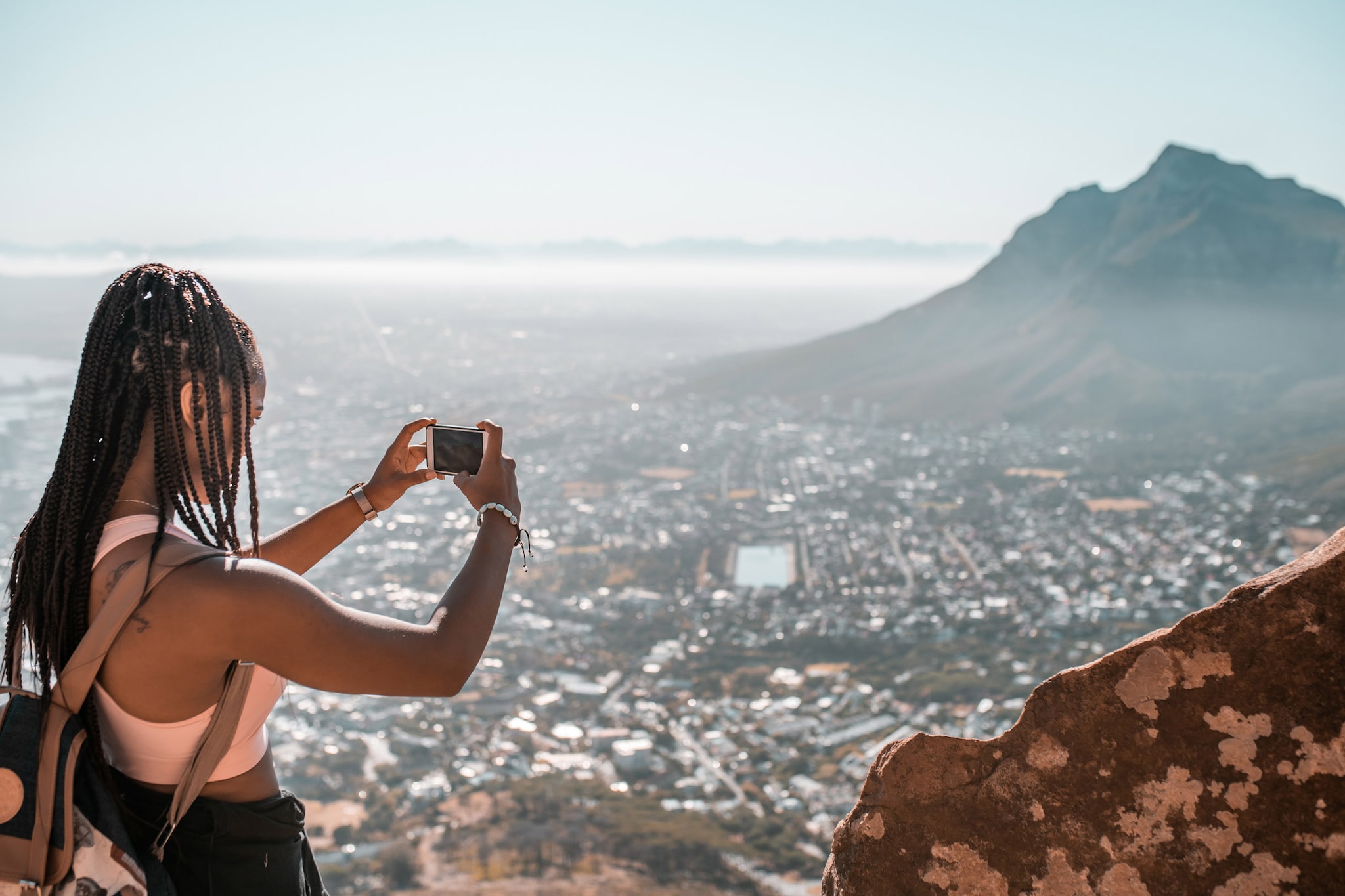 A woman stands on a mountain, taking a photo of the city below, blending nature and urban life in a stunning view.