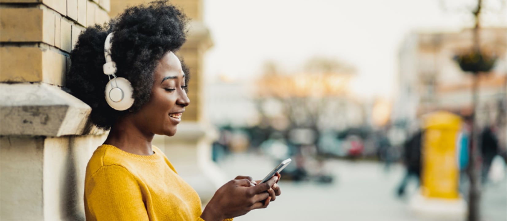 Woman is standing outside in the street with headphones on and looking through her cellphone while smiling.