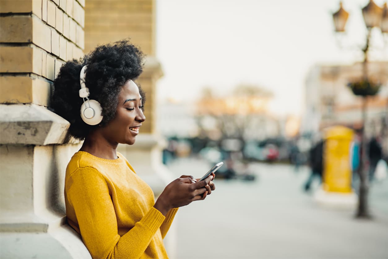 A woman leans against a wall of a building while listening to music and using her phone