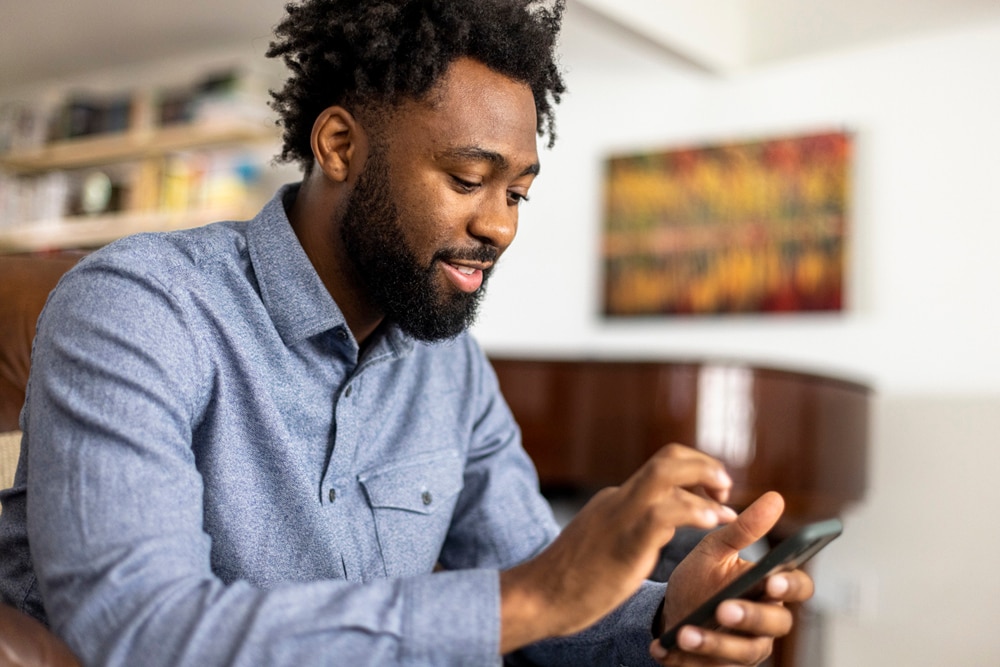 A man with a beard and laptop checks his phone, seeking guidance on beginner investment techniques.