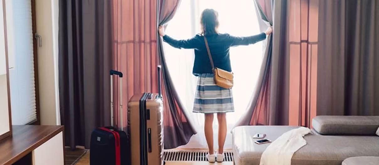 Woman looking out the window of a hotel room, holding the curtains open