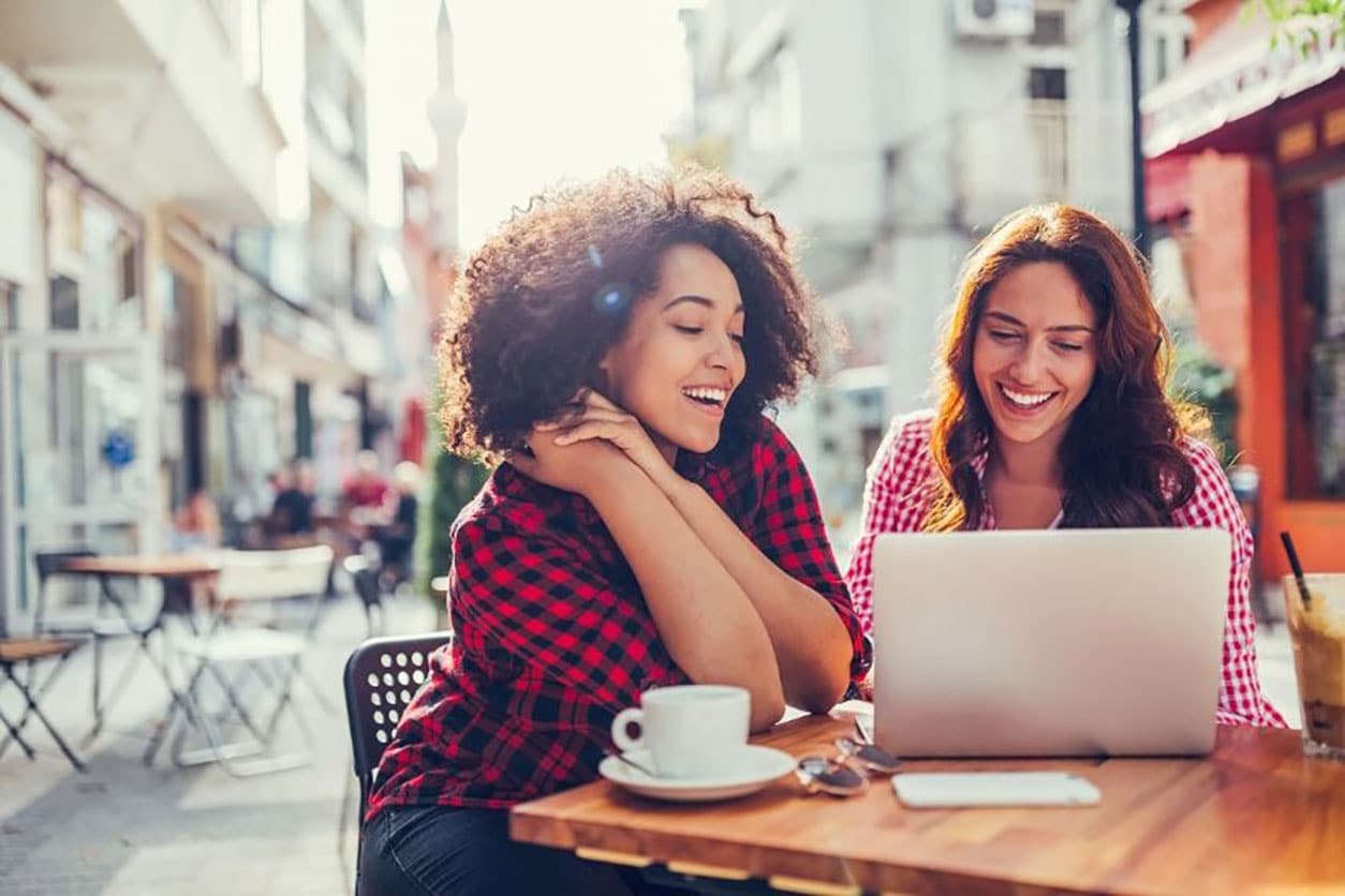 Two women are siting outside, smiling and looking at the laptop