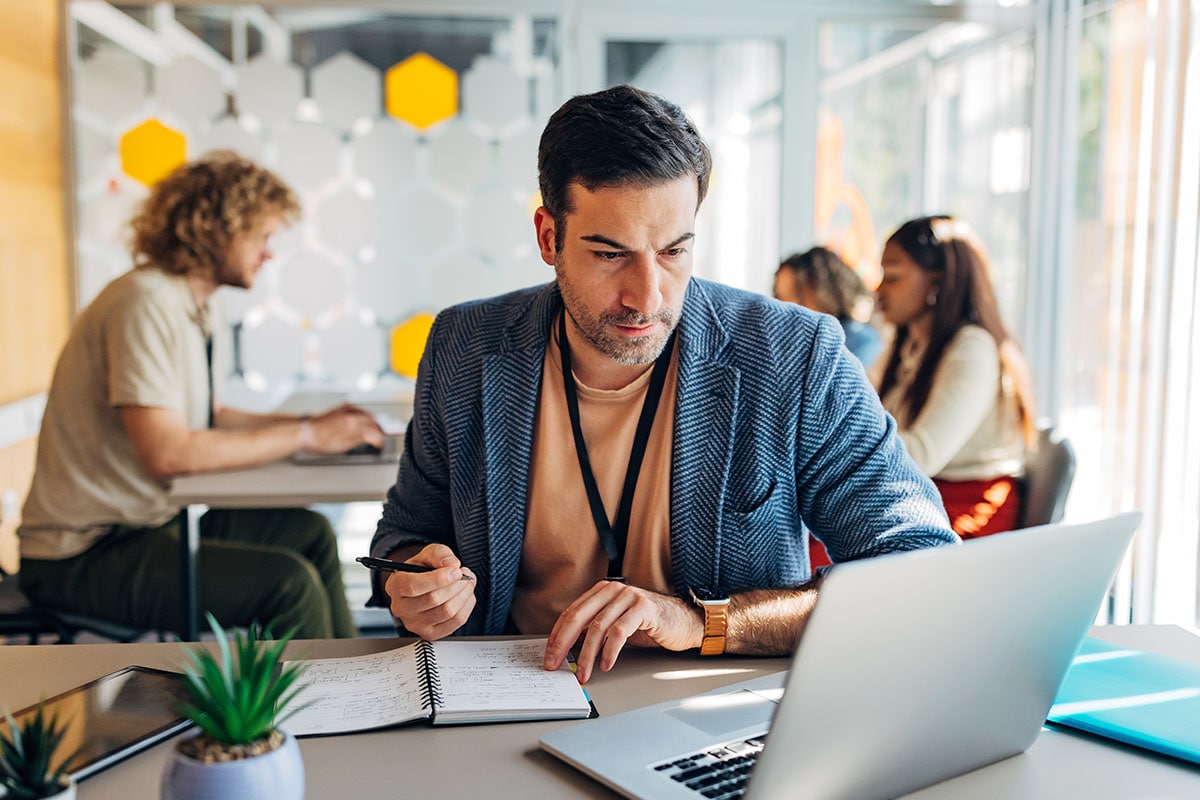Man sitting at a table with his laptop holding a pen to his notebook