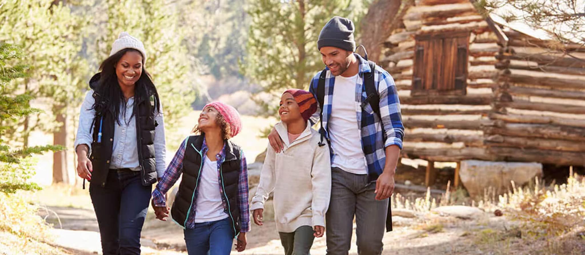 A family strolls along a scenic trail, with a cozy cabin nestled among the trees in the background.