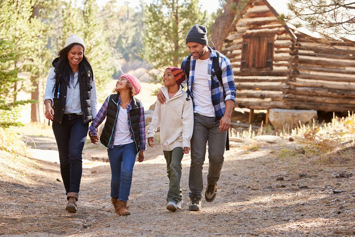 A family strolls along a scenic trail, with a cozy cabin nestled among the trees in the background.