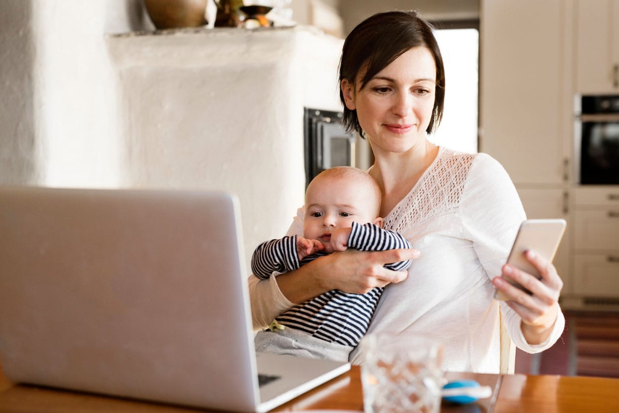 A woman sits at her dining room table while holding her baby in one hand and looking at her phone in the other