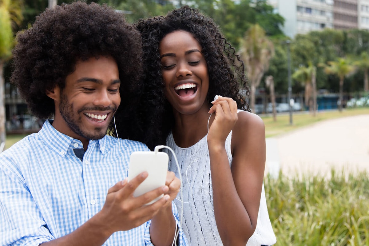 A young couple joyfully smiles and laughs together while viewing something on their cell phone, enjoying a lighthearted moment.
