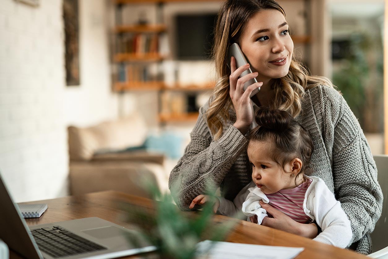 A woman holding a baby while talking on the phone with her laptop open