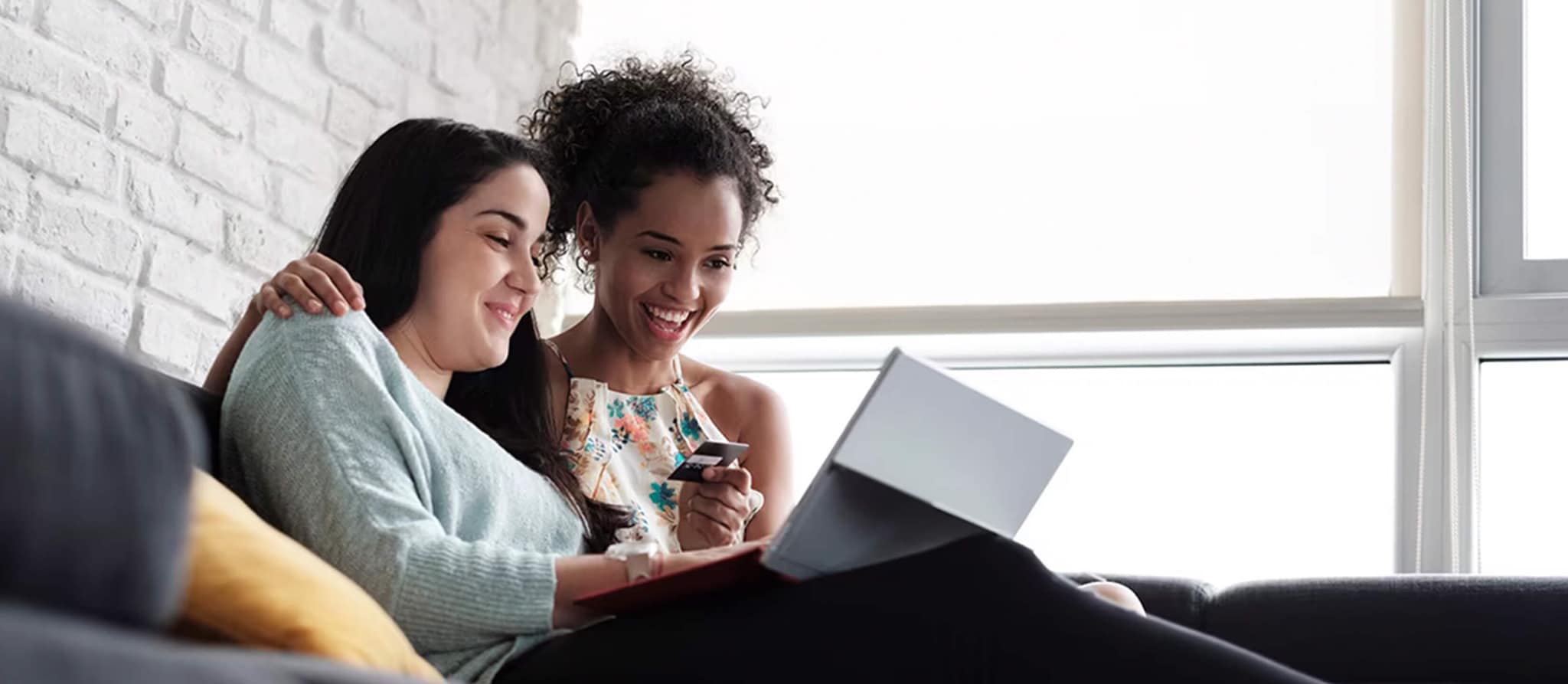 Two women seated on a couch, intently viewing a laptop screen, discussing credit card payment options.