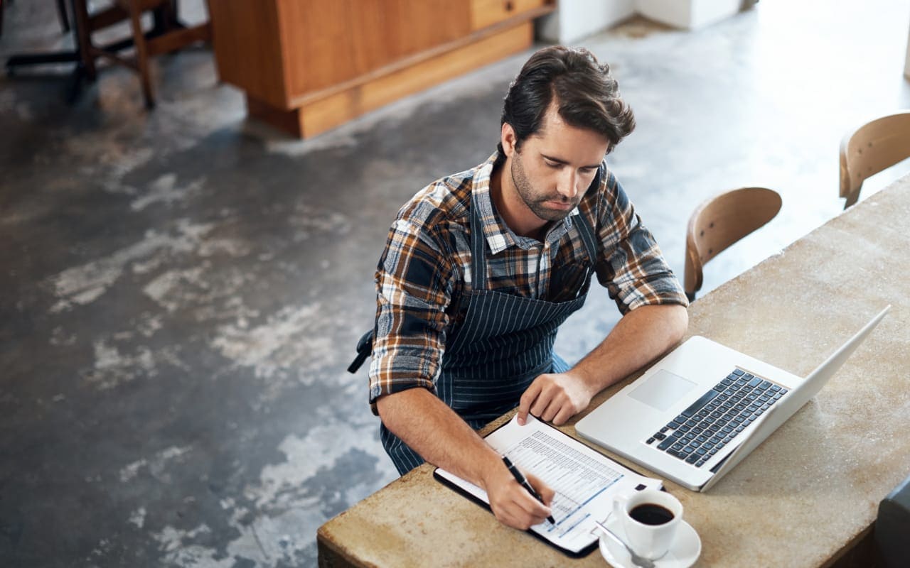 A professional image of a small business owner working on a laptop at a cafe, with a notebook and coffee nearby, evoking productivity and financial management.