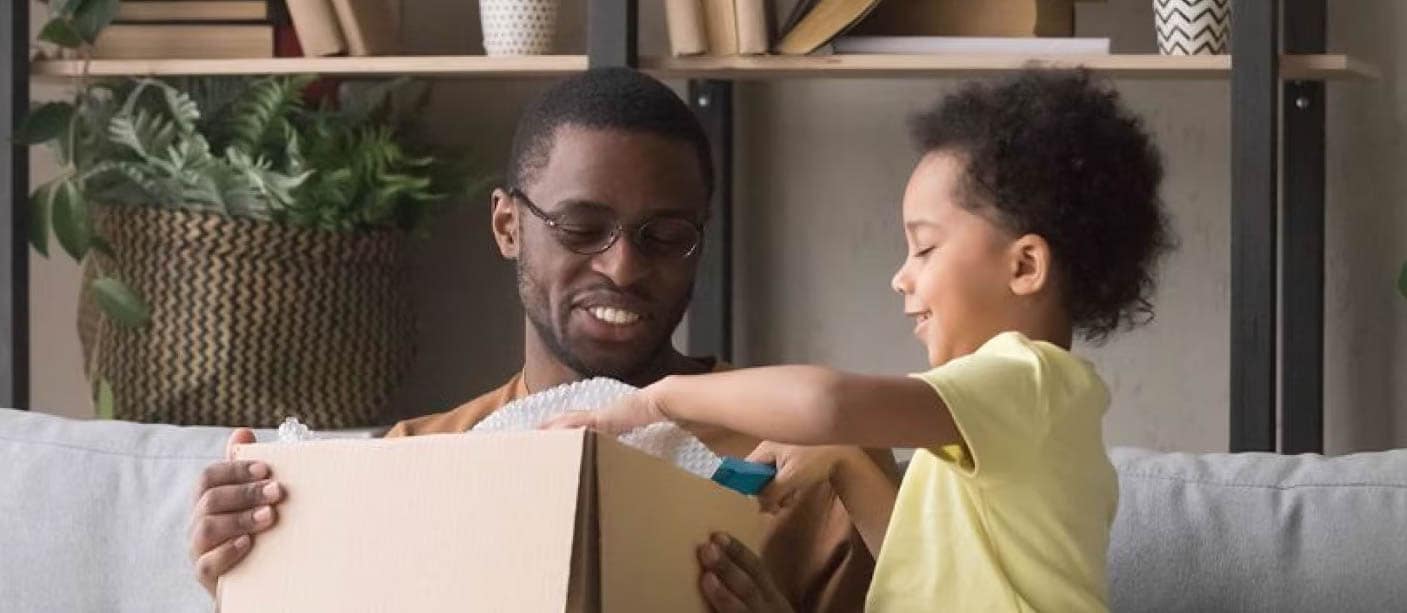 A man smiling while sitting on a couch holding an open box and a young child smiling looking through the contents