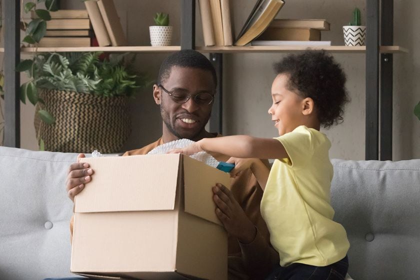 A man smiling while sitting on a couch holding an open box and a young child smiling looking through the contents