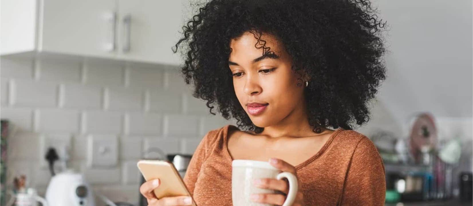 A woman checks her loan balance via her phone while drinking coffee in her kitchen