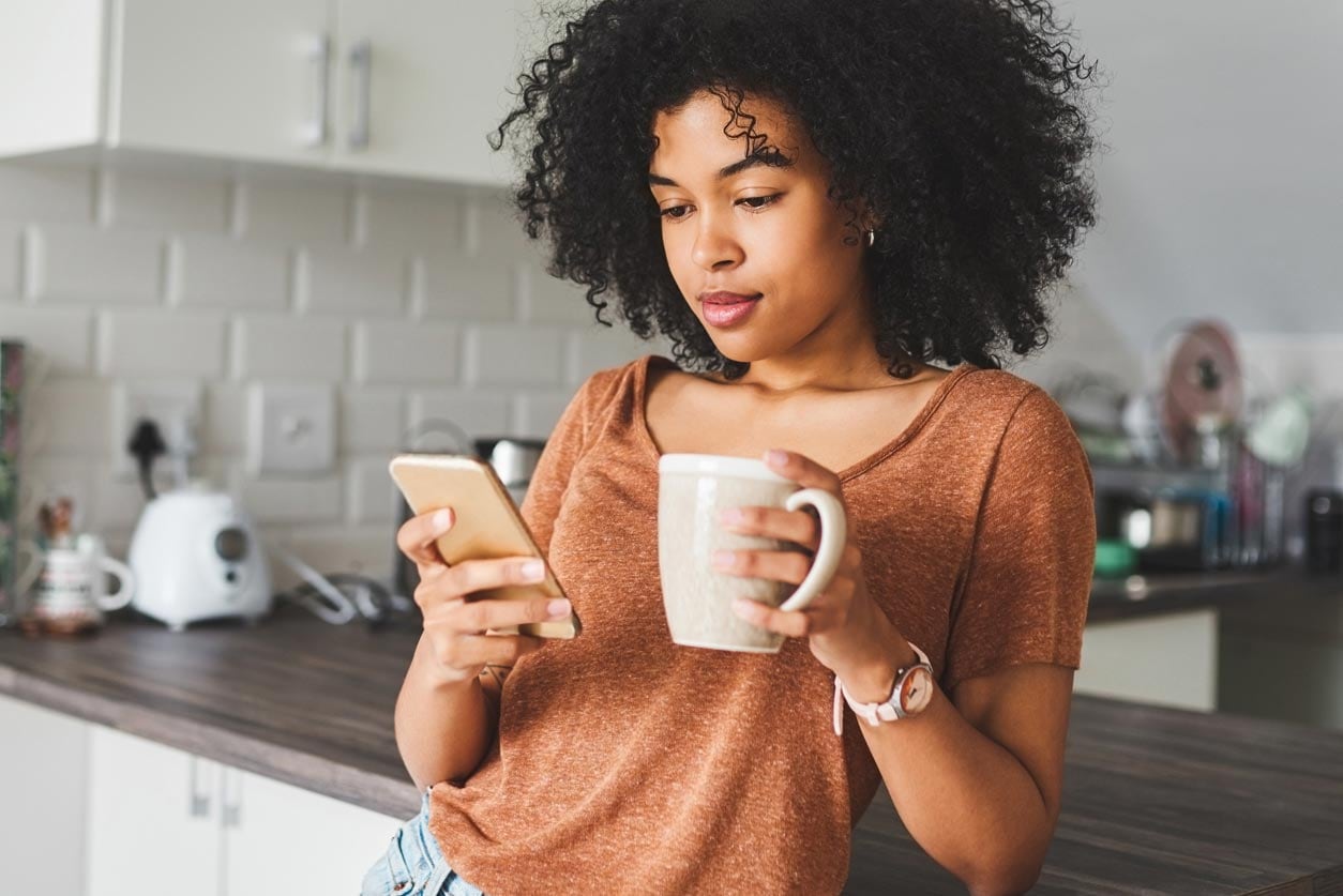 A woman checks her loan balance via her phone while drinking coffee in her kitchen