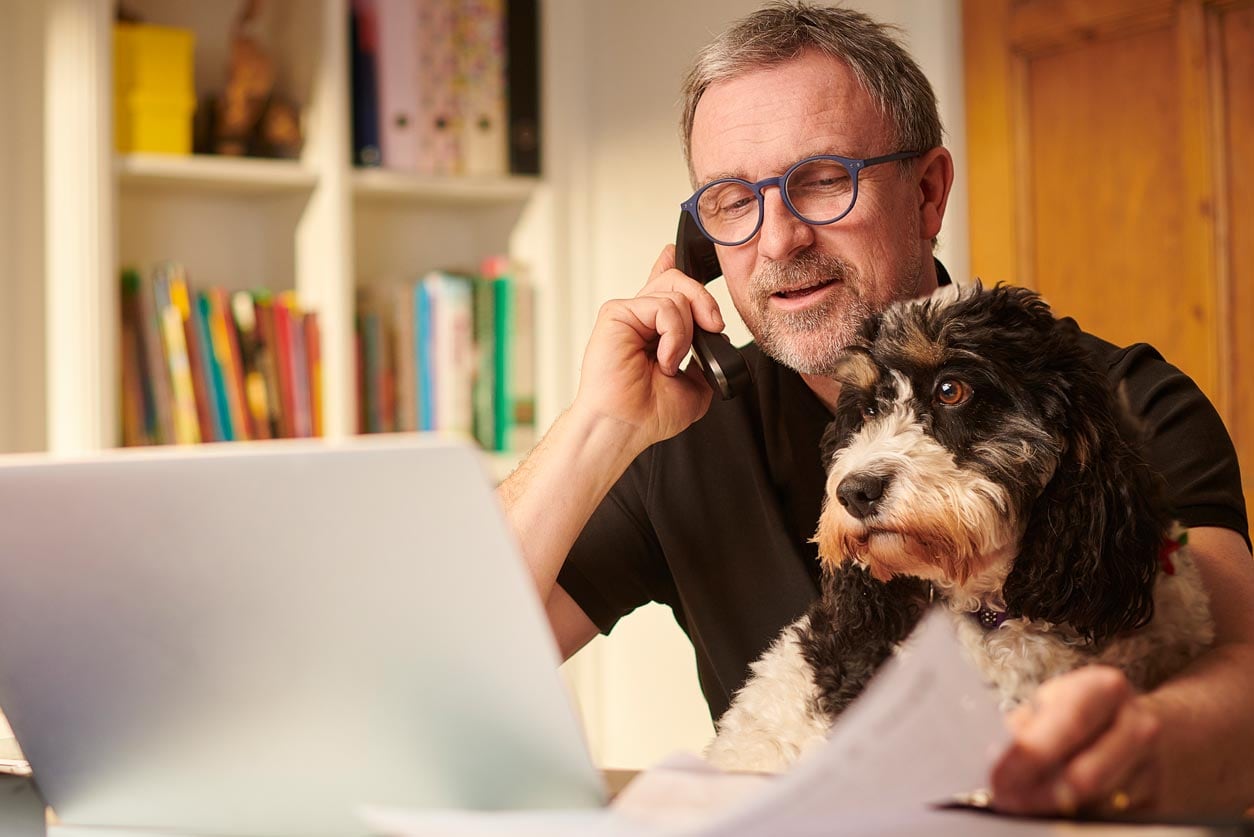 A man is on the phone going through papers and looking at his laptop with his dog on his lap.