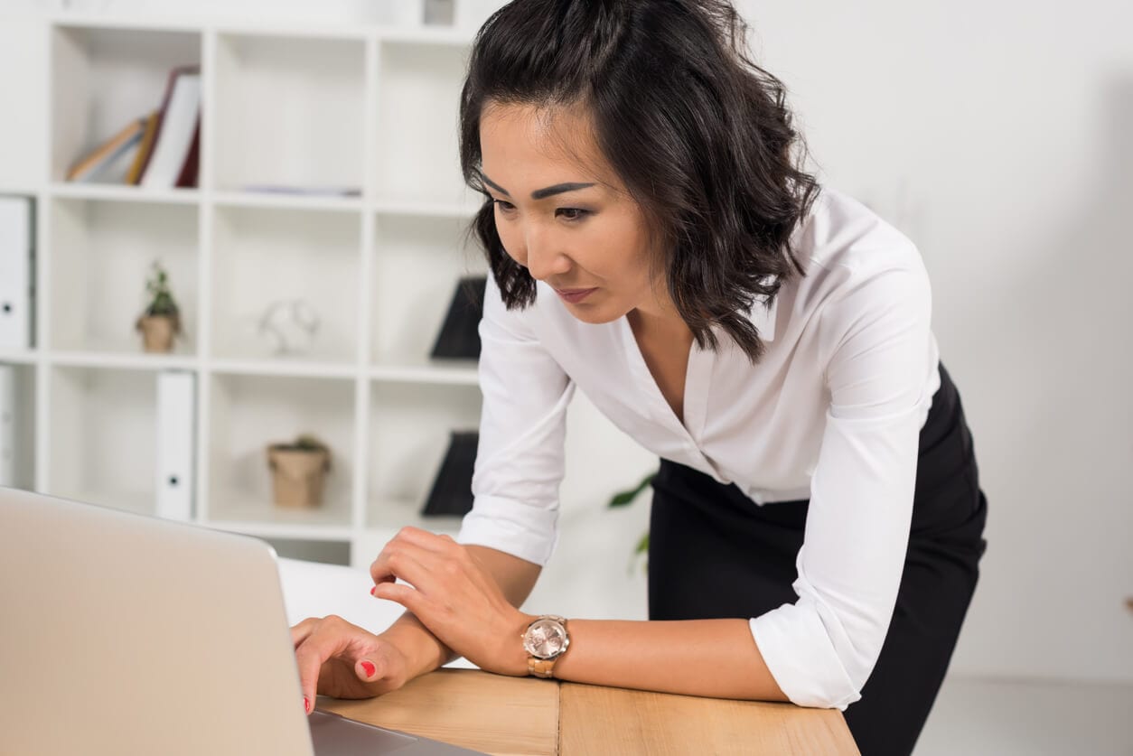 woman standing and leaning on a desk while working on a laptop