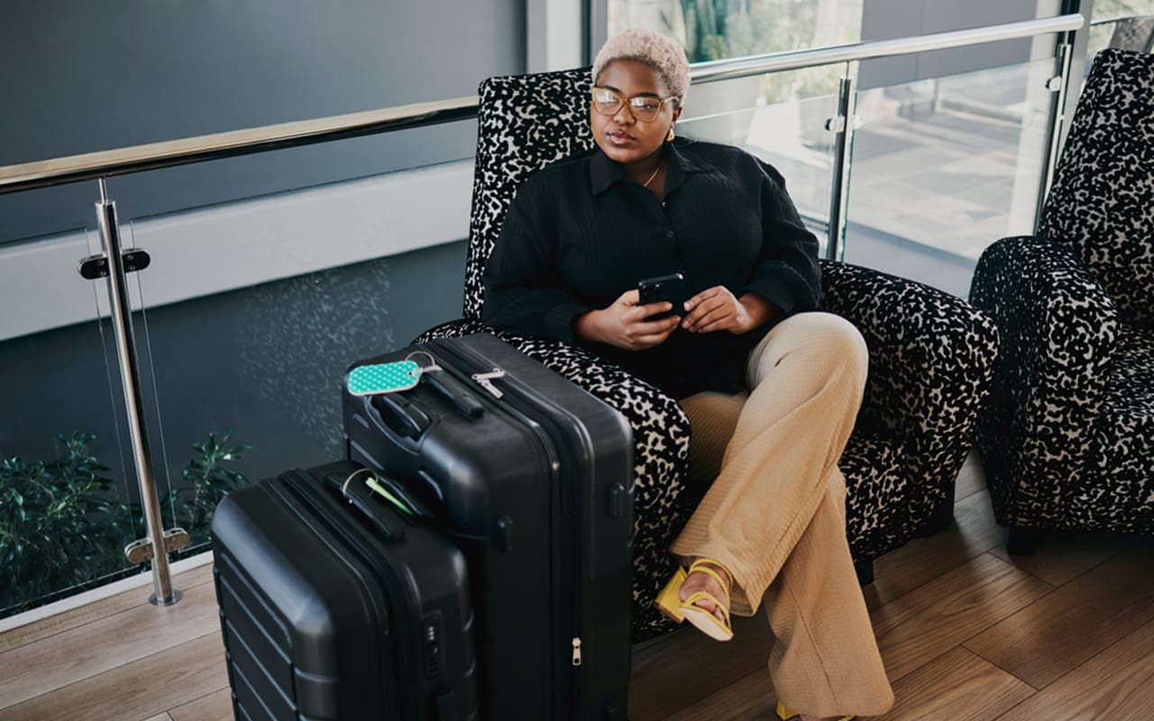 A lifestyle photo of a young traveler at an airport lounge, smiling while holding a passport and boarding pass, with a suitcase nearby.