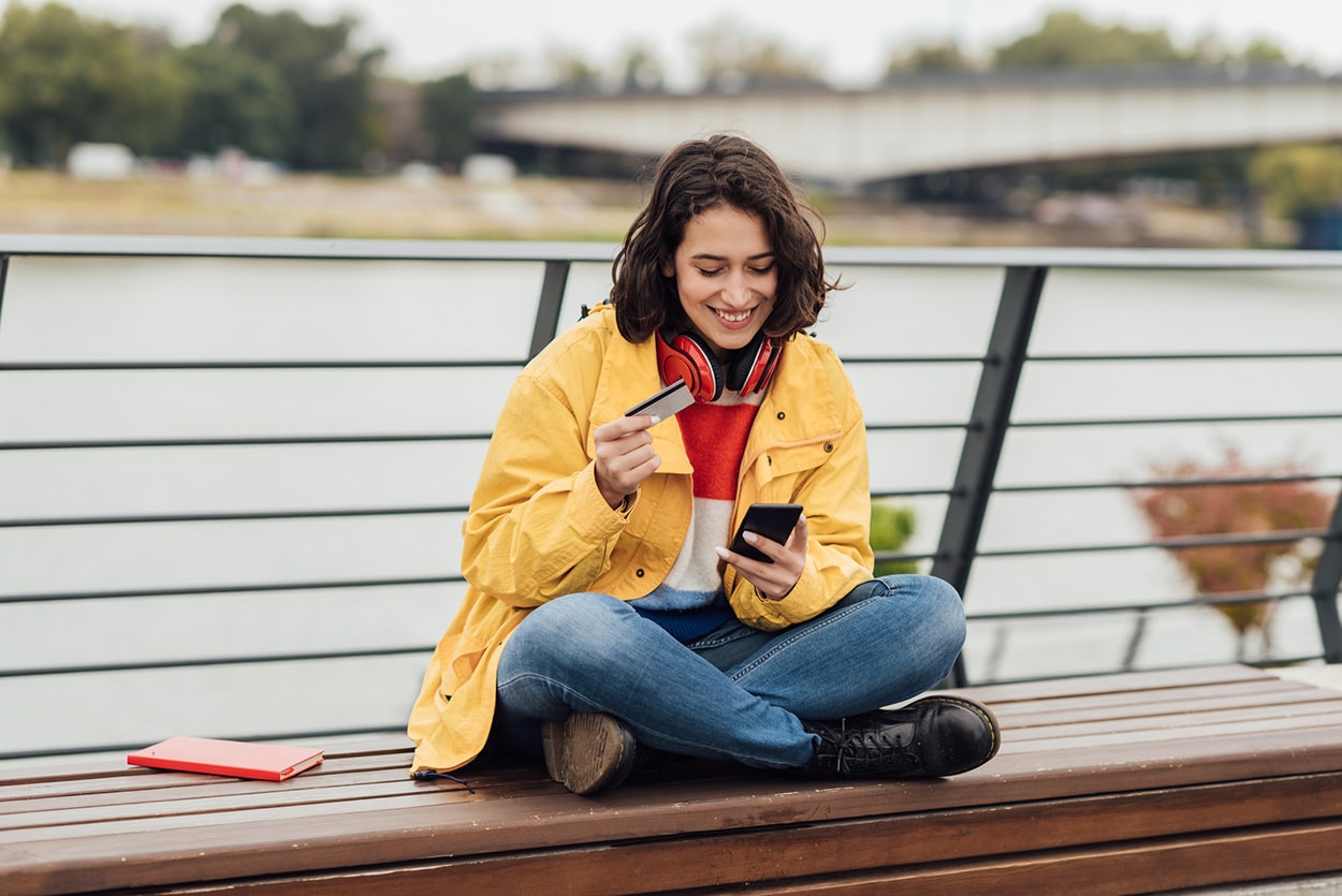 A teen sits on a boardwalk by a lake looking at her phone while holding her credit card