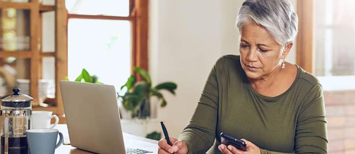 older woman is sitting at a table in front of a laptop and writing down on a paper while holding a cellphone in other hand 