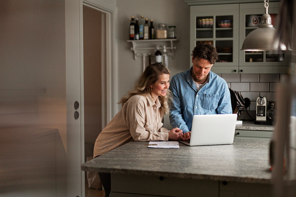 A bright lifestyle photo of a focused traveler sitting at a kitchen island, reviewing booking documents and a passport next to an open laptop and a cup of coffee.