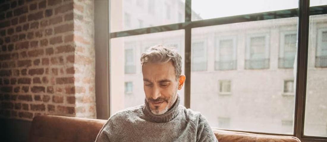 A man sits on a couch with a laptop, contemplating the pros and cons of withdrawing retirement funds early.