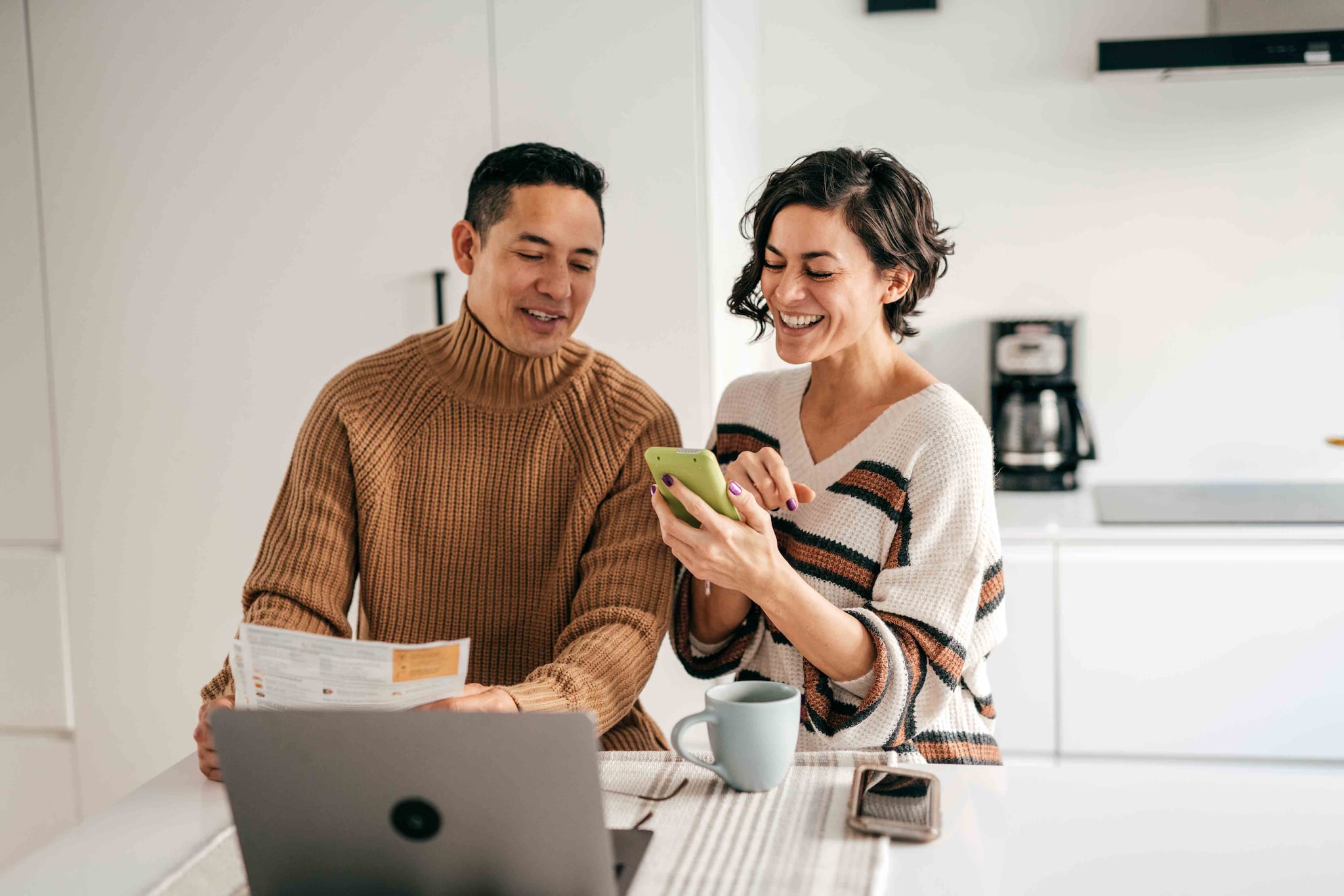 Couple sitting at a kitchen island, looking at a phone with papers and a laptop in front of them