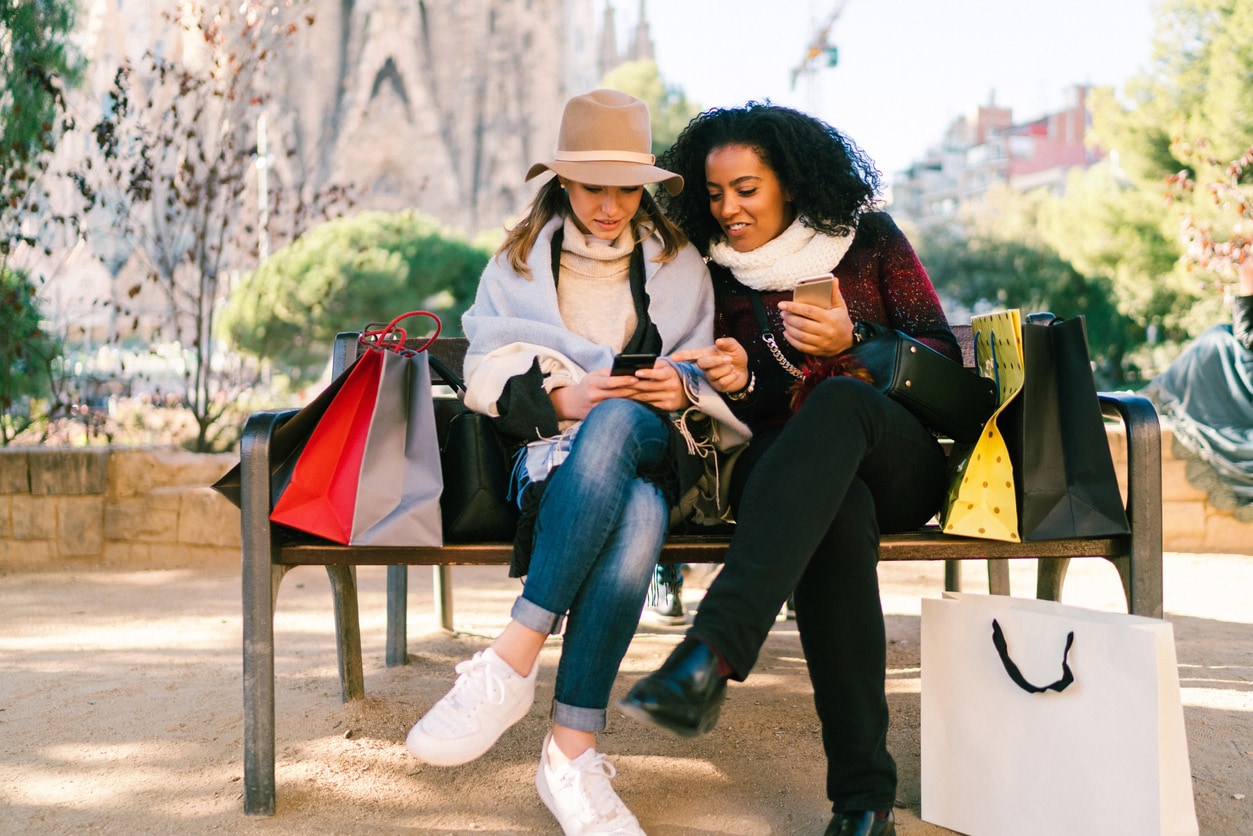 Two girls are sitting on a bench in a park looking at their cellphones
