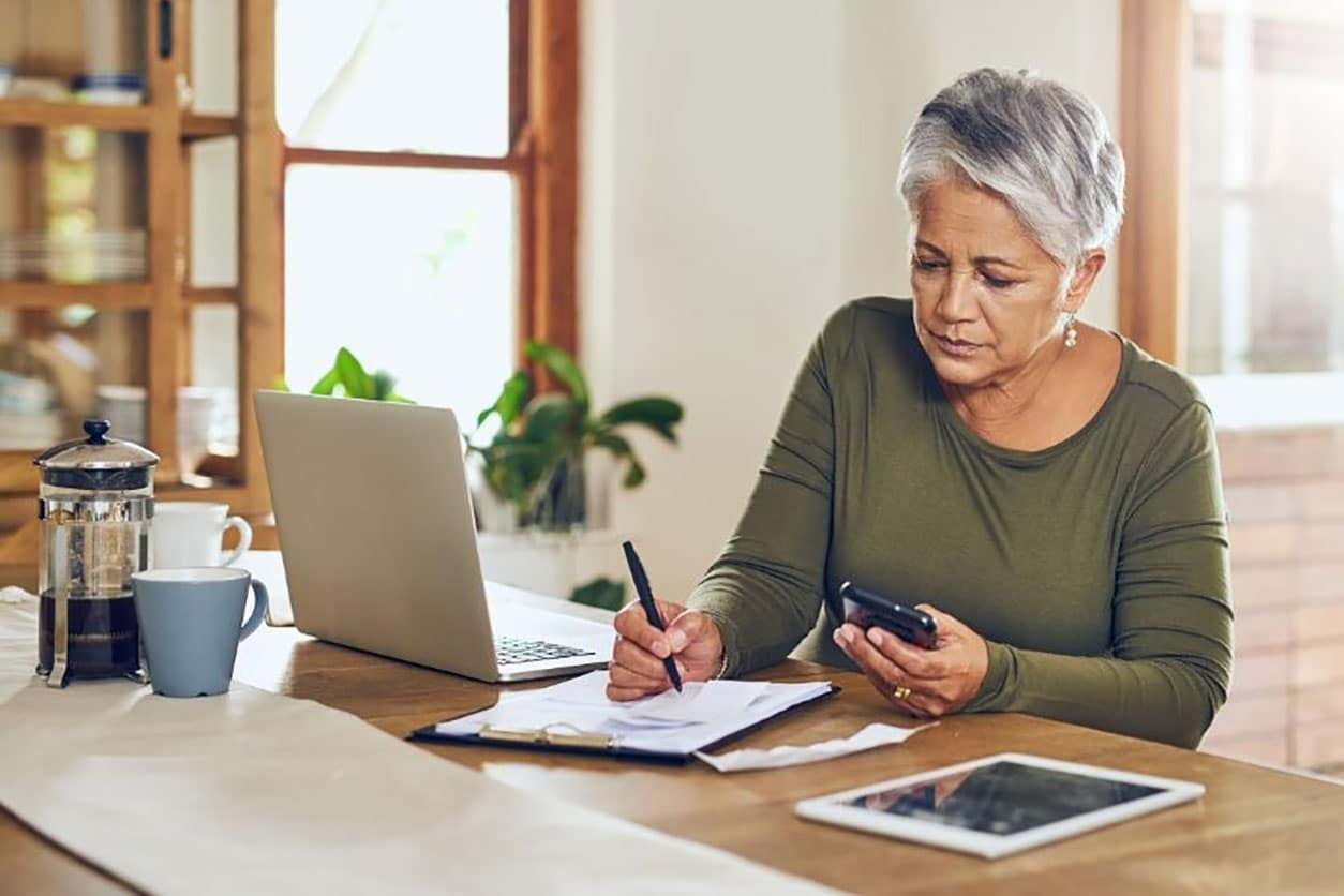 older woman is sitting at a table in front of a laptop and writing down on a paper while holding a cellphone in other hand 