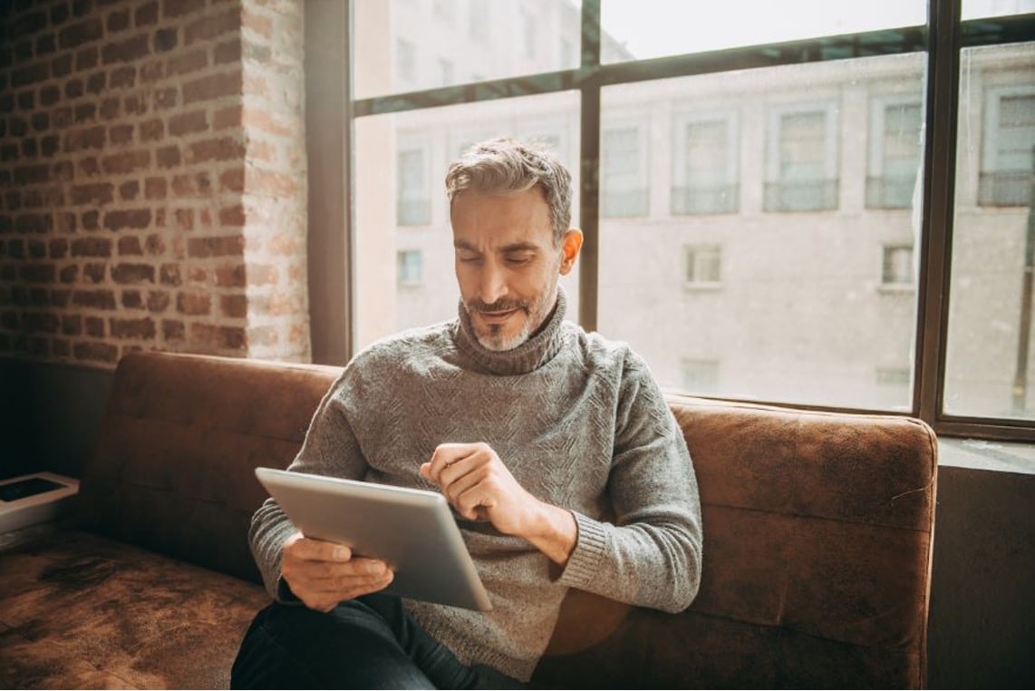 A man sits on a couch with a laptop, contemplating the pros and cons of withdrawing retirement funds early.