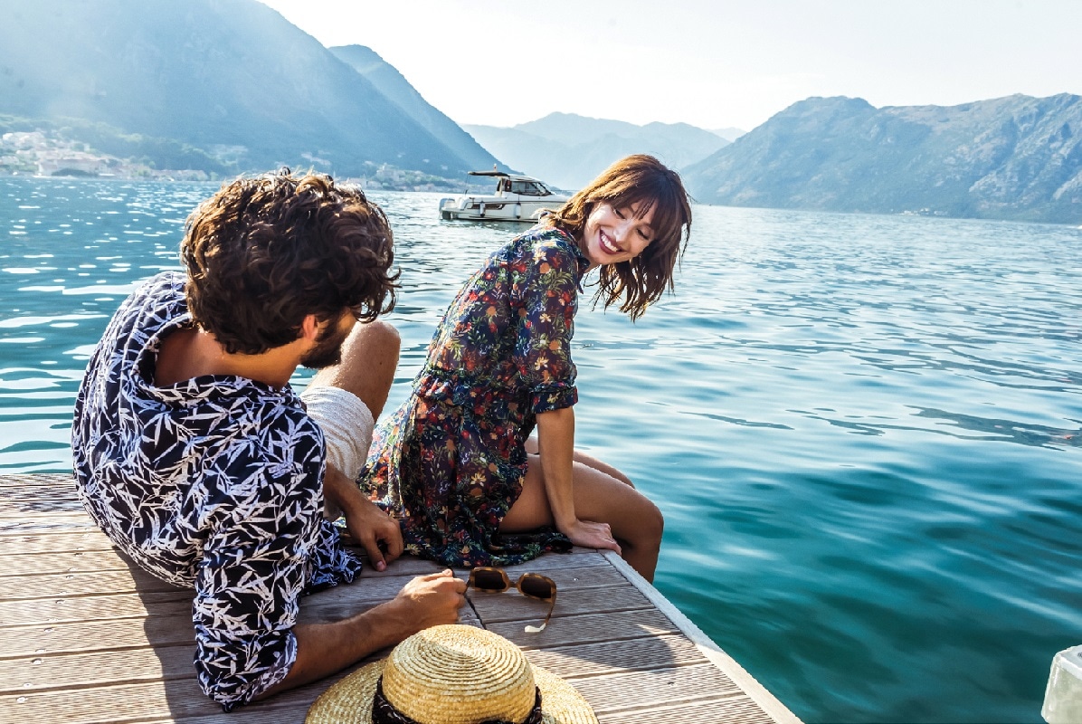 Two people sitting the edge of a boat dock by a lake laughing with mountains in the background