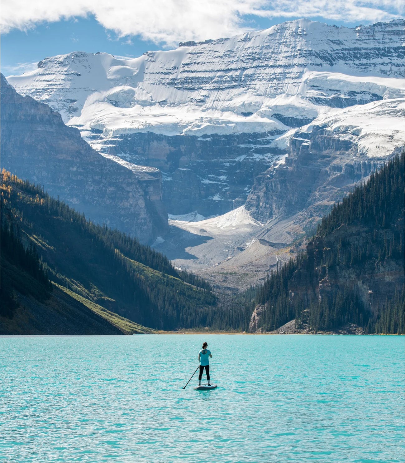 Una persona practica paddle surf en un lago de aguas turquesas rodeado de imponentes montañas nevadas y bosques de pinos.