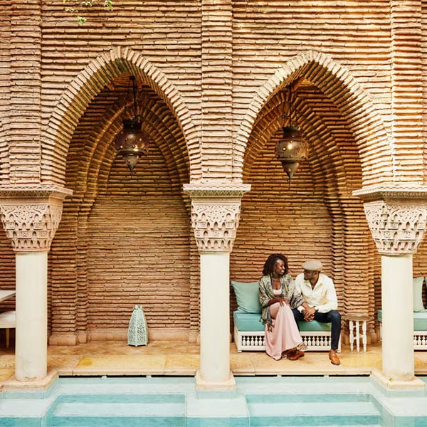 Una pareja conversa sonriente sentada en un sofá bajo arcos de ladrillo de estilo árabe con lámparas colgantes decorativas.