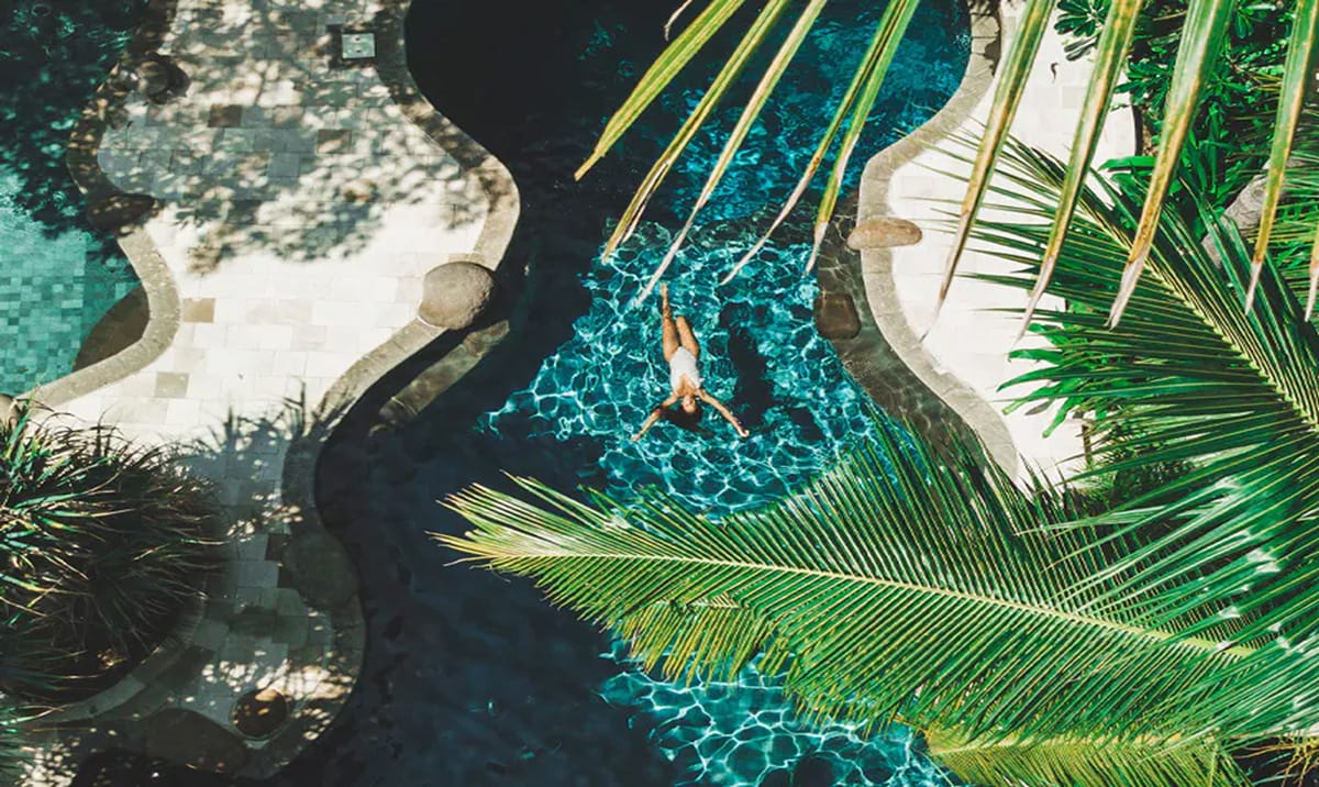 Vista aérea de una persona nadando boca arriba en una piscina de aguas cristalinas rodeada de vegetación tropical y palmeras.