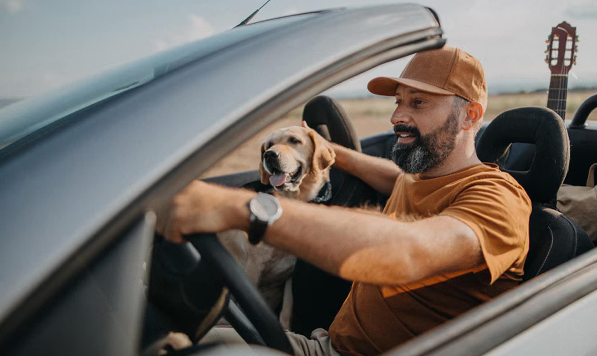 Un hombre con barba y gorra conduce un coche descapotable junto a su perro labrador dorado en el asiento
del copiloto.