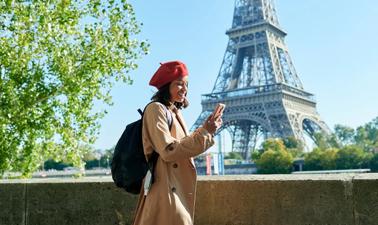 Una joven sonriente con boina roja y gabardina mira su teléfono móvil con
la Torre Eiffel de fondo en París.