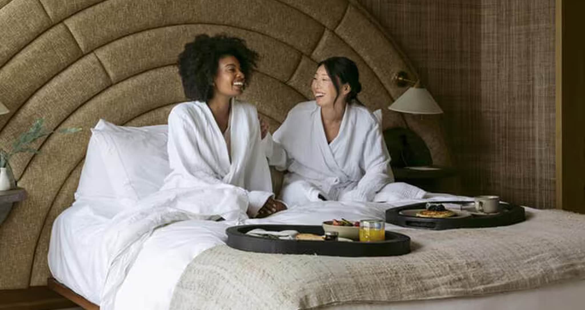 Dos mujeres con batas de baño blancas ríen felices sentadas en una cama
grande con bandejas de desayuno servidas.