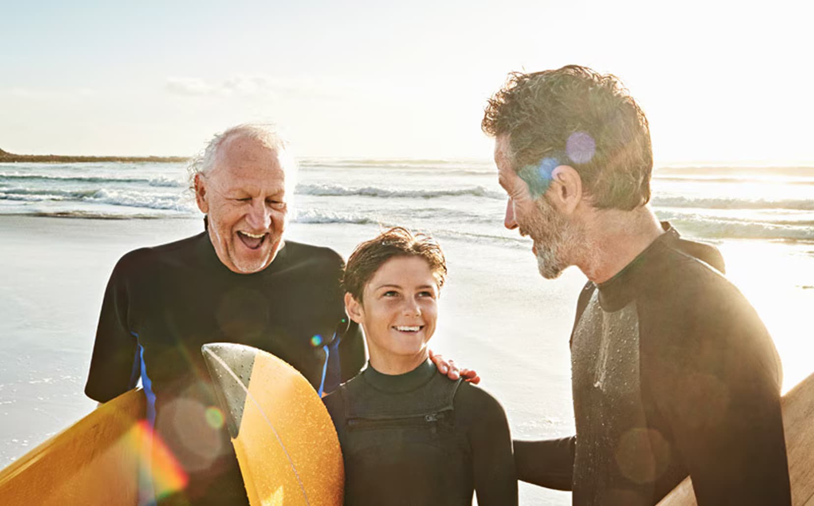 Tres surfistas de distintas generaciones con trajes de neopreno ríen juntos en la playa sosteniendo sus tablas de surf al atardecer.