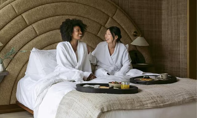 Dos mujeres con albornoces blancos sentadas en una cama de hotel, riendo juntas con bandejas de desayuno servidas frente a ellas.