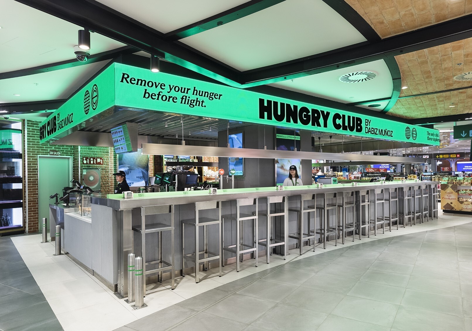 Barra y mostrador del restaurante Hungry Club por Dabiz Muñoz, con un gran letrero luminoso de color verde brillante y personal trabajando al fondo.
