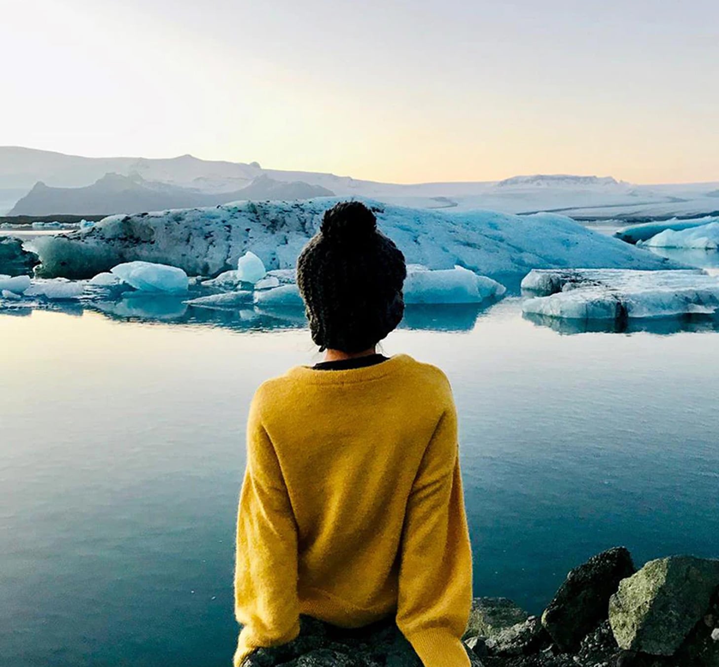 Una persona de espaldas con un suéter amarillo y un gorro de lana observa un lago glacial con icebergs flotando y montañas al fondo durante el amanecer o atardecer.