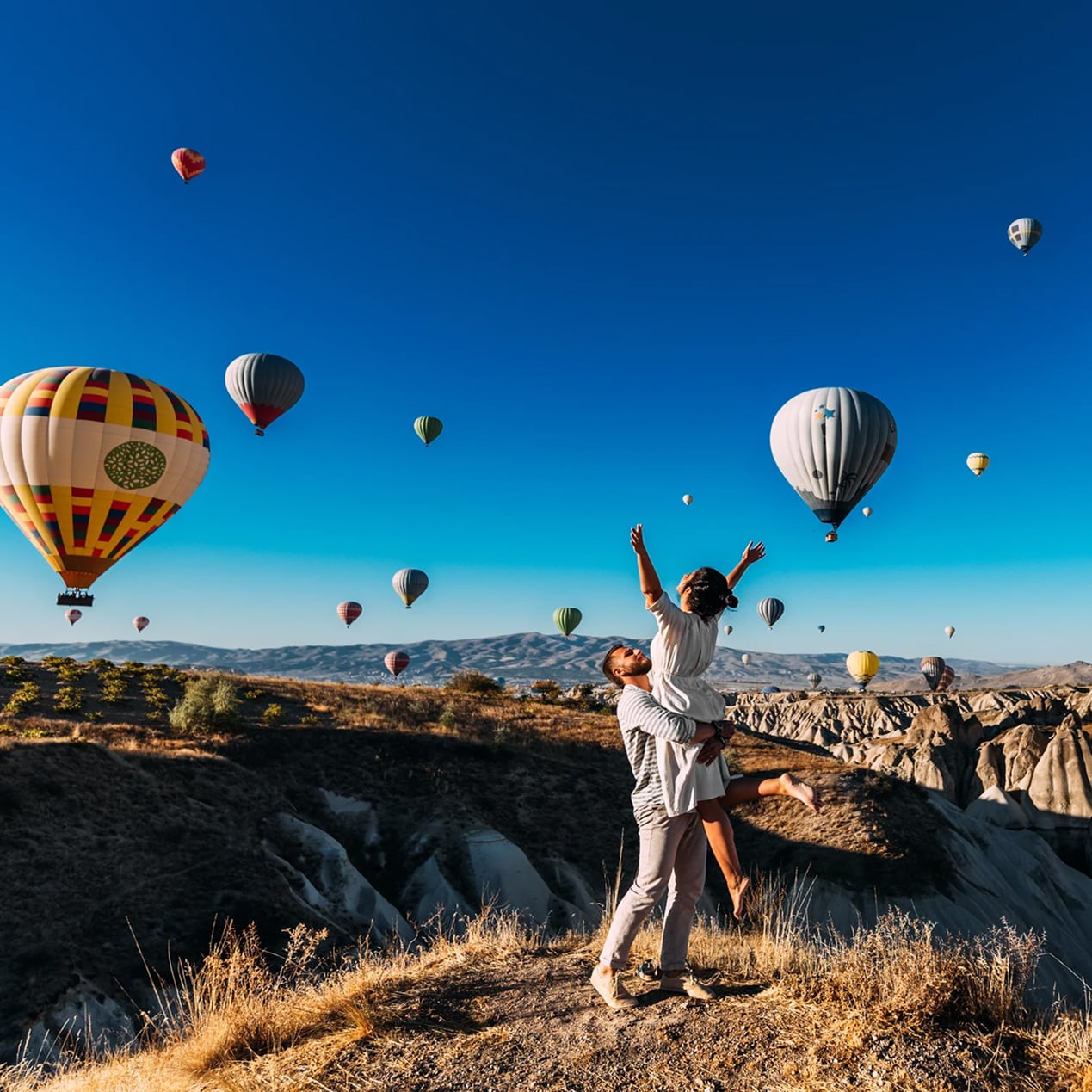 Pareja abrazándose en una colina de Capadocia con globos aerostáticos en el cielo azul.