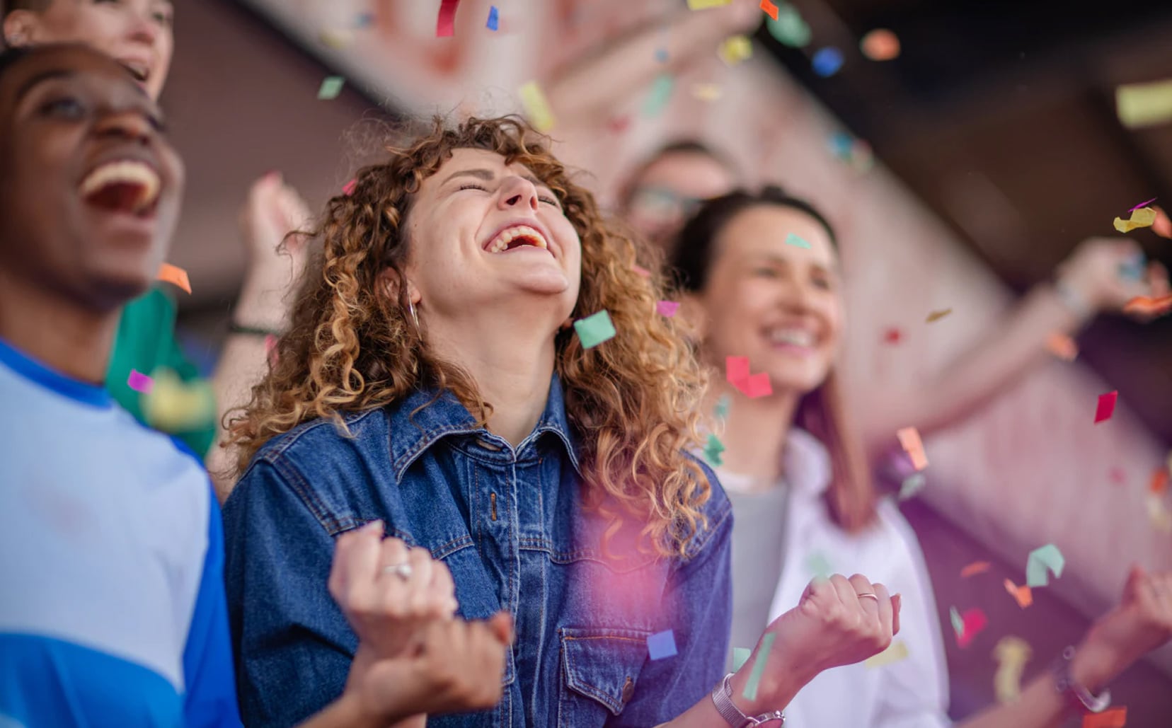 Un grupo de personas celebra con entusiasmo en una tribuna mientras cae confeti de colores, destacando en primer plano una mujer con cabello rizado y chaqueta de mezclilla que ríe con los puños cerrados.