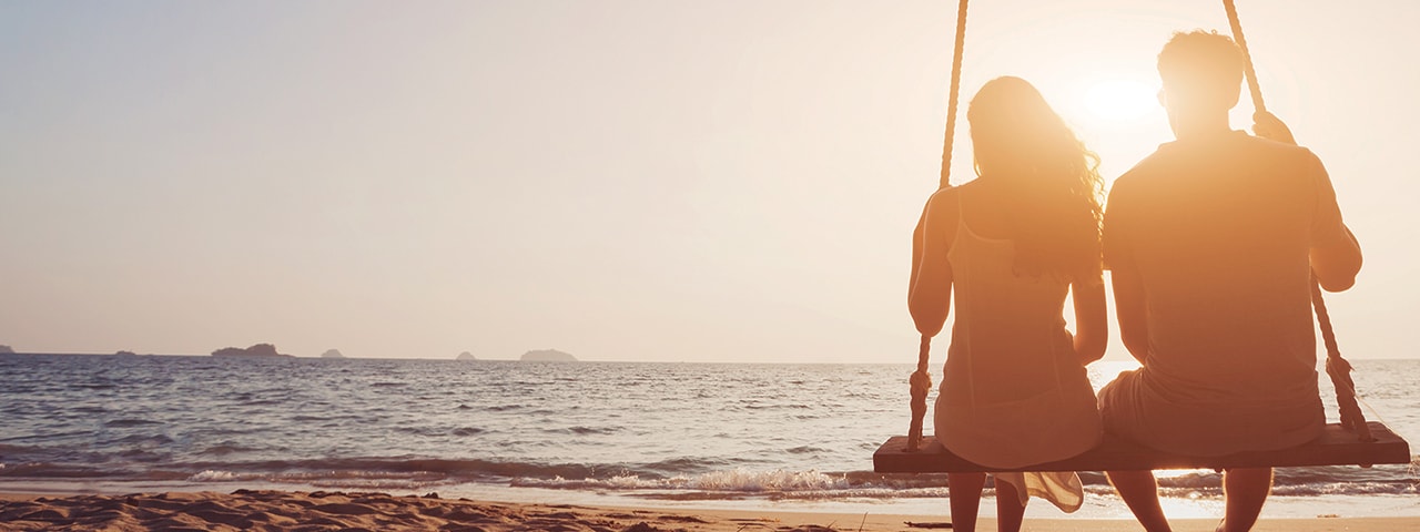 Una pareja en un columpio en la playa mirando la puesta de sol.