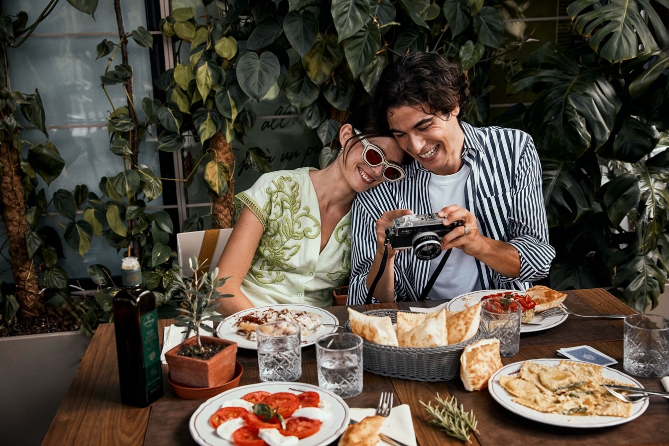 Personas sentadas del mismo lado enfrente de una mesa con comida, y ambos están sonriendo hacia una cámara – AMEX MX
