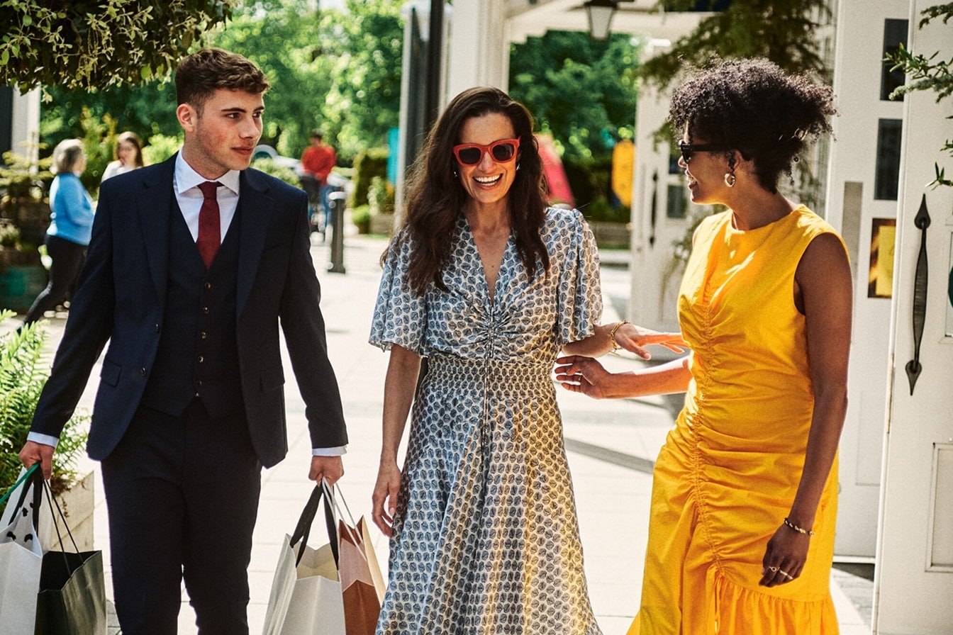 Dos mujeres riendo y caminando, mientras que hay un hombre con traje al lado cargando bolsas de compras – AMEX MX