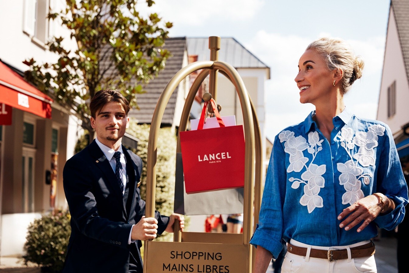 Una mujer sonriendo, y detrás hay un hombre de traje moviendo un carrito de equipaje con bolsas de compras – AMEX MX 
