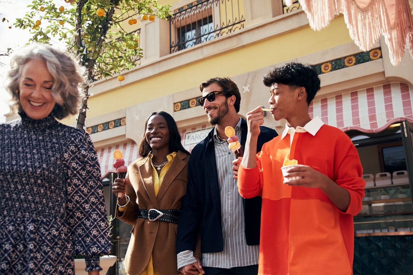 Una mujer caminando al frente sonriendo, mientras que detrás hay una mujer y dos hombres riendo y comiendo helado – AMEX MX