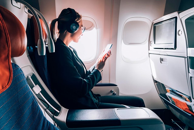 Una mujer sentada en un avión, con auriculares y mirando su teléfono.