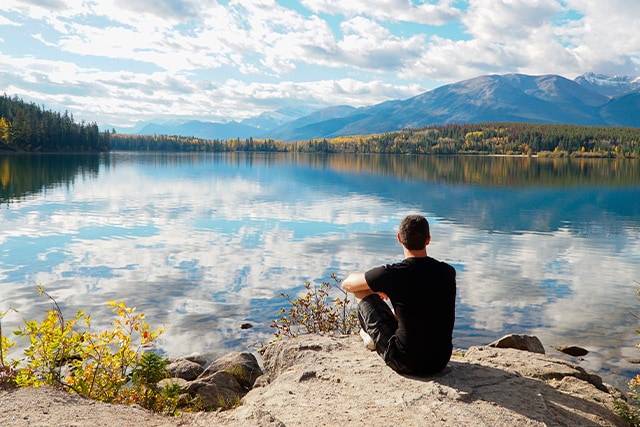Un hombre sentado solo sobre una roca, con vistas a un lago y un paisaje montañoso.